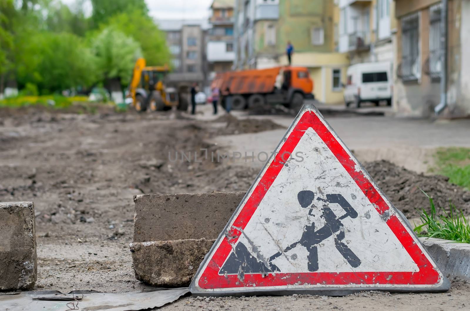 sign road repair stands on the background of the destroyed asphalt pavement in the courtyard of a multi-storey building, construction equipment and workers