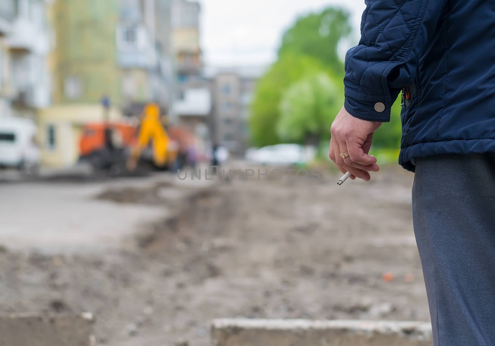 Hand man with a cigarette on the background of the destroyed and repair the road in the yard of a house with construction machinery and machines