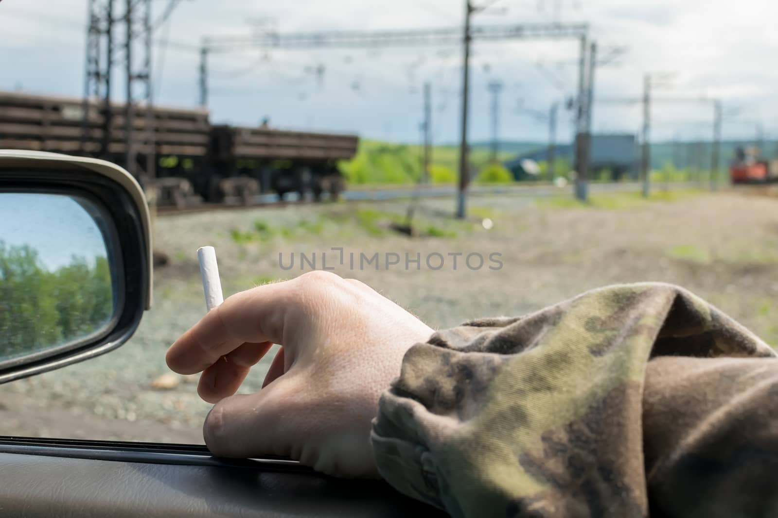 hand men, military, in camouflage clothing, holding a cigarette while in the car, which is near the railway crossing with freight cars