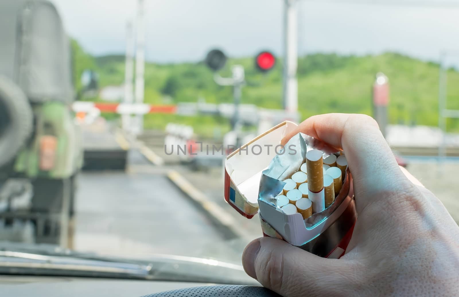 View of the driver hand with a pack of cigarettes on the steering wheel of the car, which stopped before a closed railway crossing at a red light