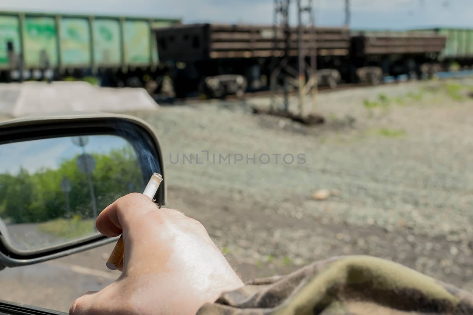 hand men, military, in camouflage clothing, holding a cigarette while in the car, which is near the railway crossing with freight cars