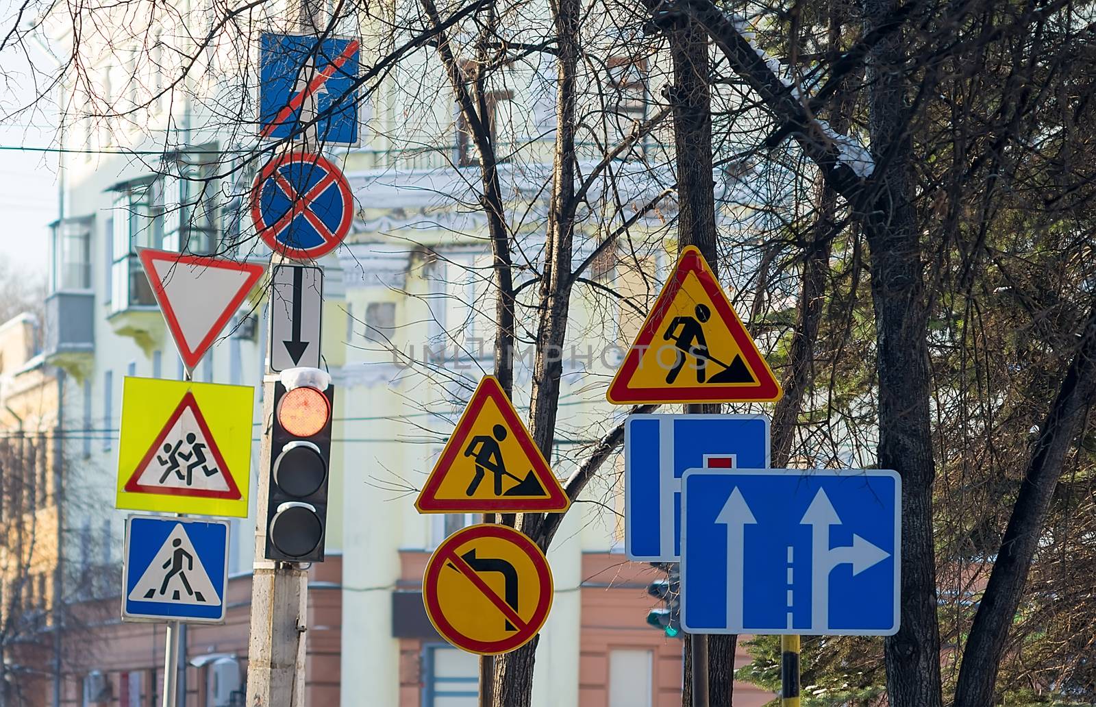 a large number of road signs, symbols of detours, road repairs near the traffic light with a pedestrian crossing on the background of city buildings and tree branches