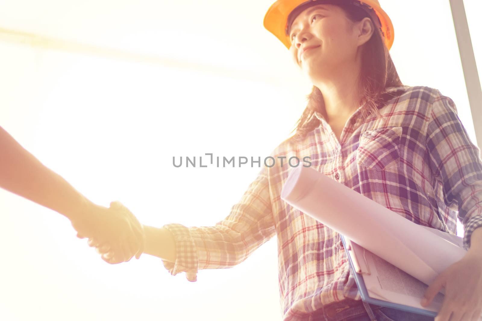 happy Asian woman engineer as construction workers with blueprint and contract in hand with protective yellow helmet are shaking hands while working at construction site by asiandelight