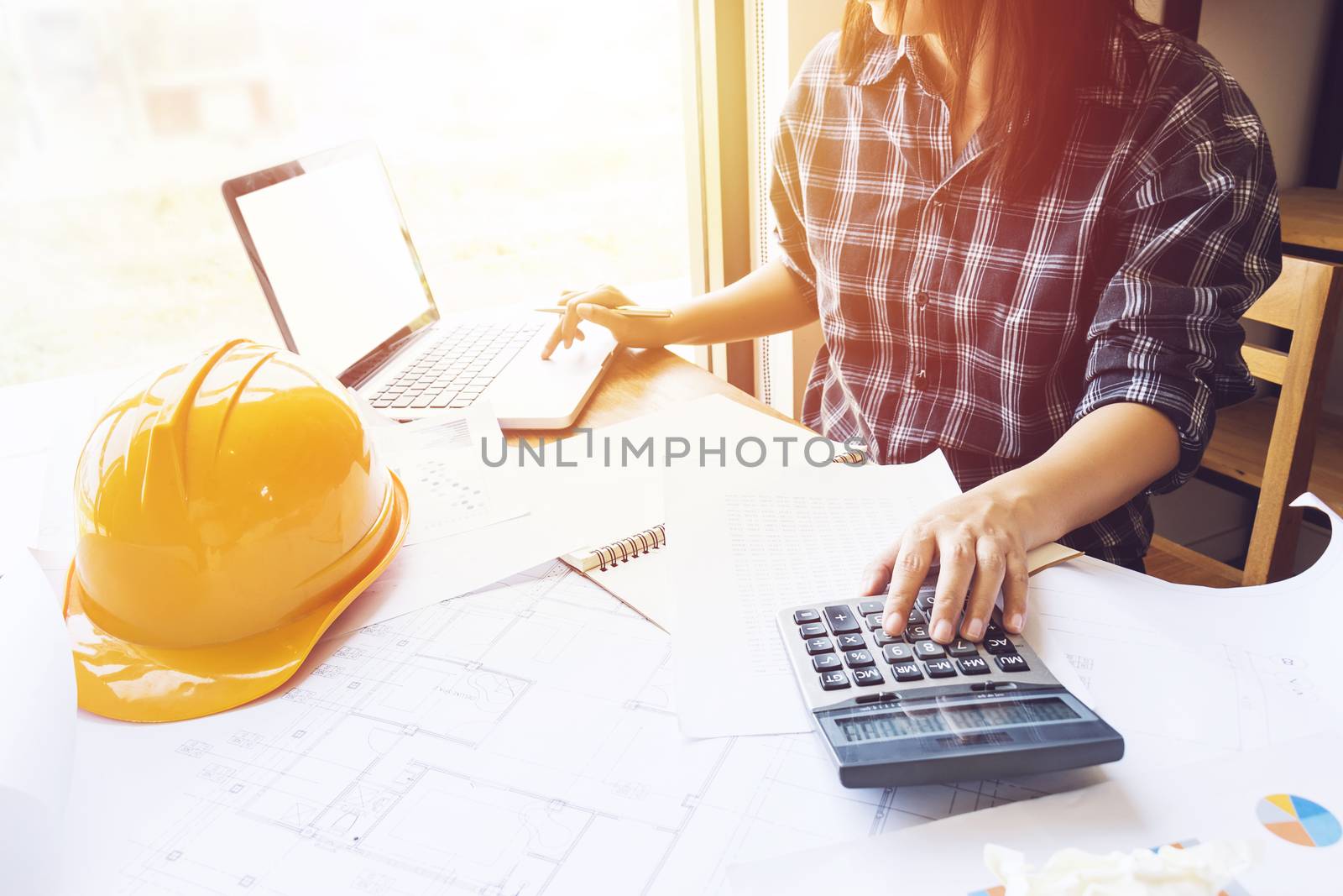 Asian engineer , architect woman using calculator for accounting and analyzing building structure from blueprint and paper chart in front of computer laptop at office work space.