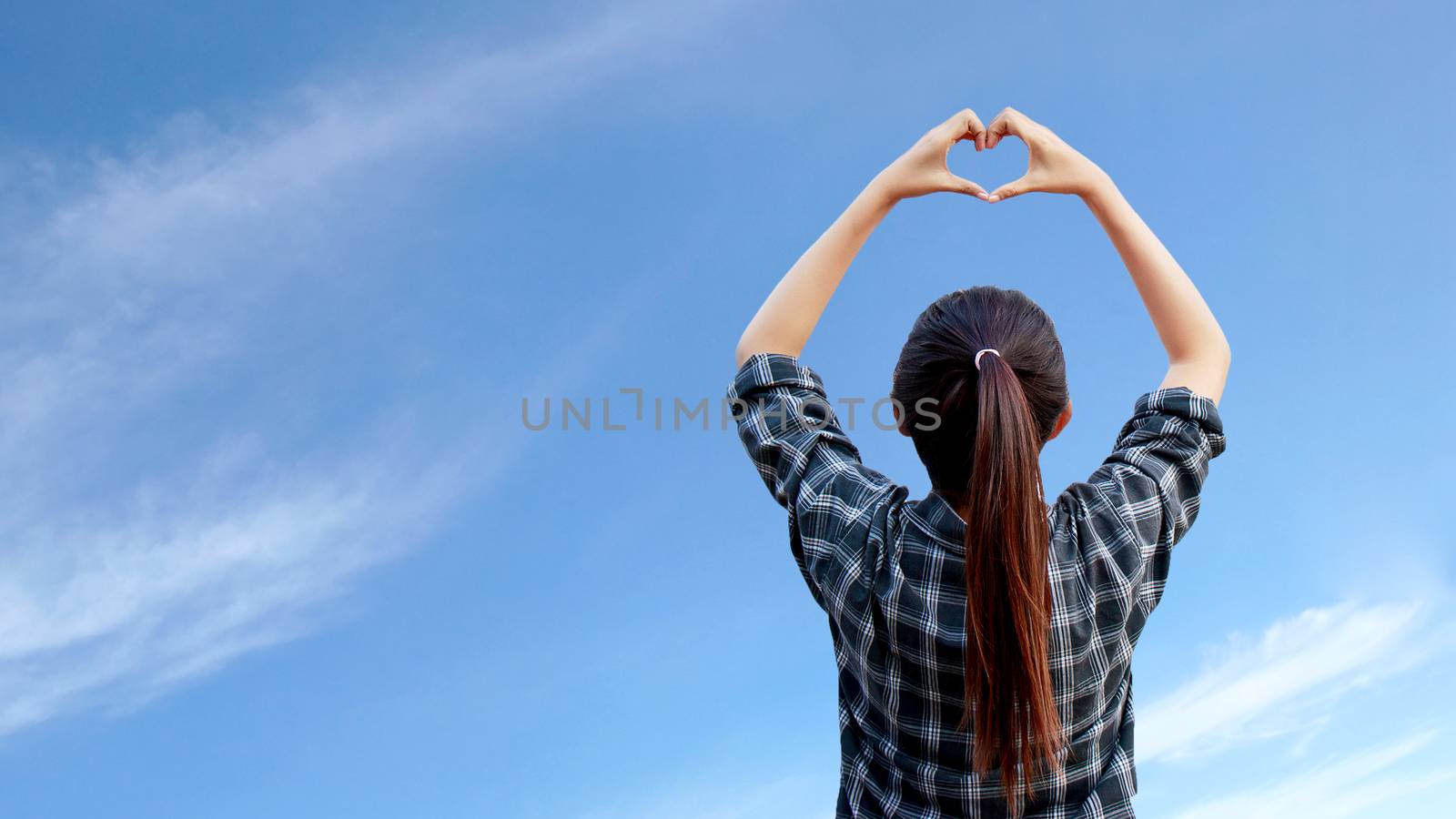 woman raise her hands to make heart shape in the air with blue sky at background by asiandelight