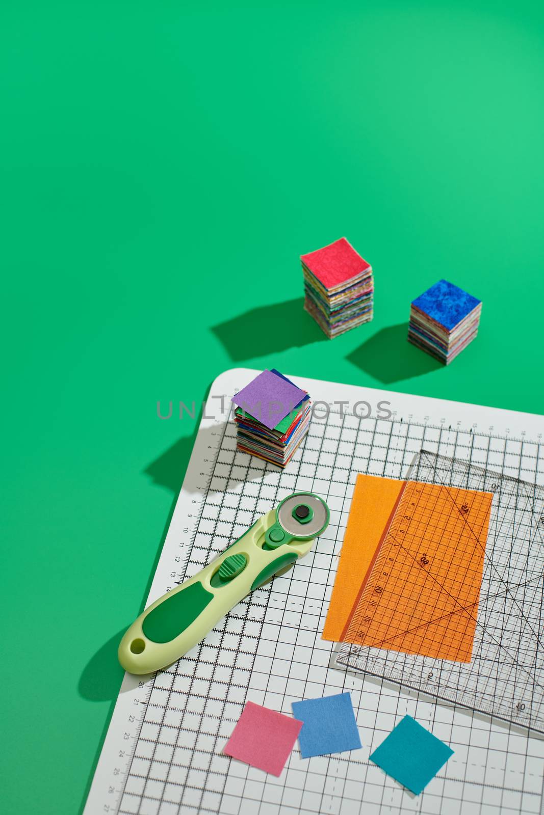 Rotary cutter, ruler, bright square pieces of fabric, stack of bright square fabric pieces on craft mat, green background