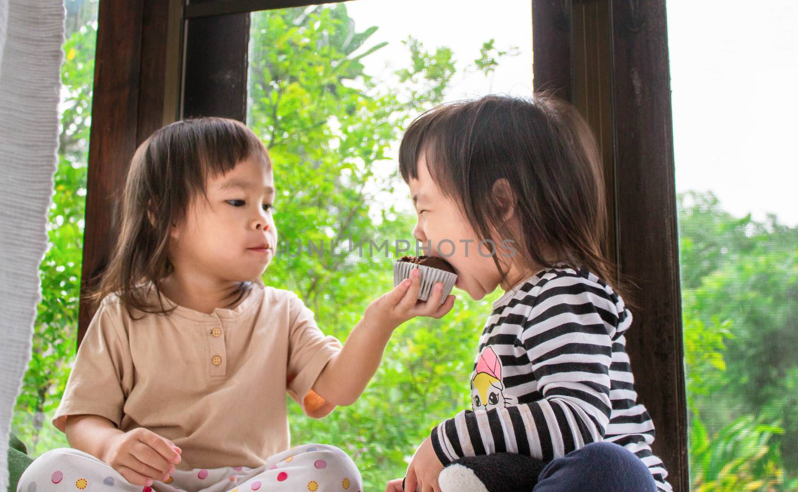 Asian little child girl and her sister are eating banana cupcake in the room deliciously. by TEERASAK