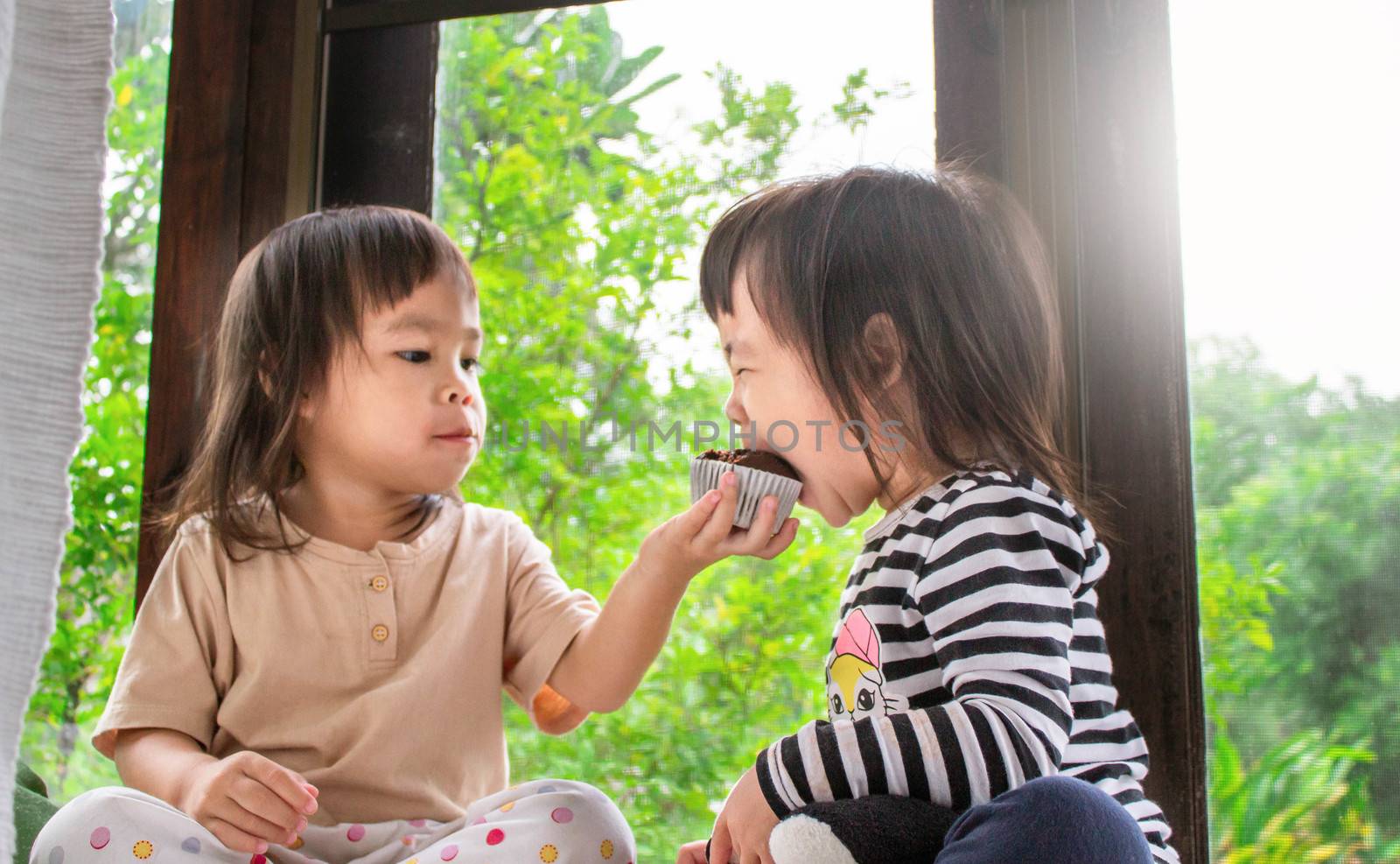 Asian little child girl and her sister are eating banana cupcake in the room deliciously.