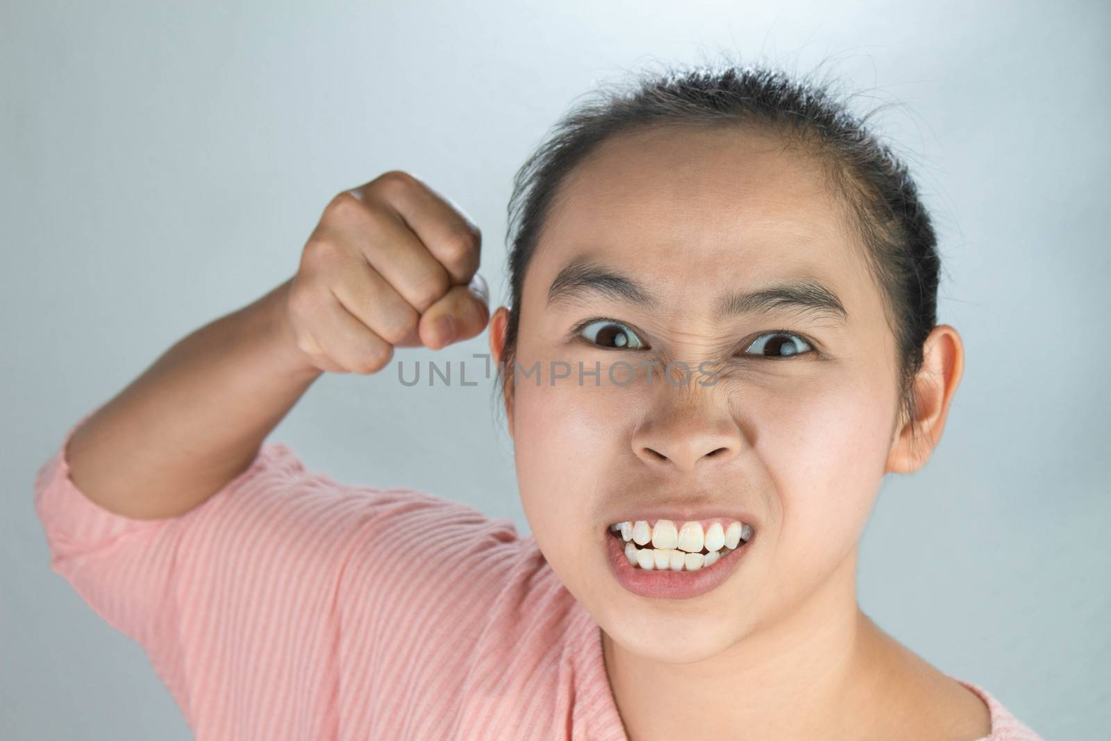 Portrait angry face of Asian young woman in pink shirt and raising fist frustrated on grey background.