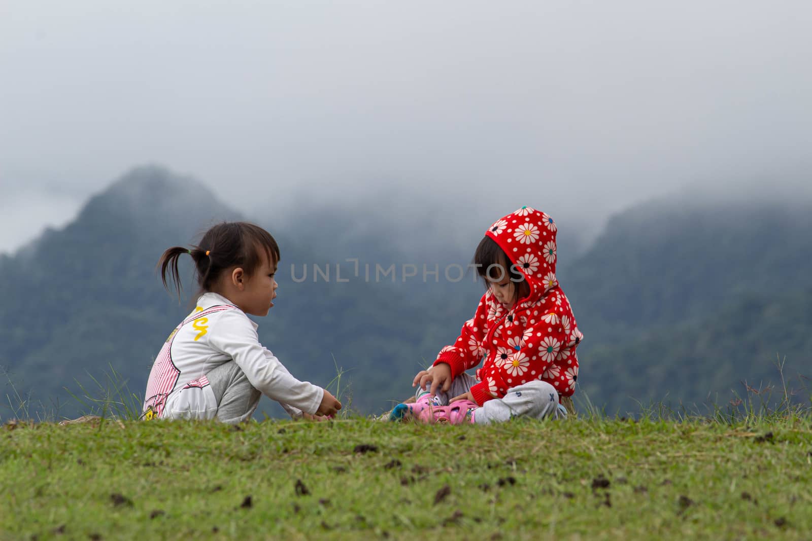 Asian little child girl helped to put shoes on the sister on the grass at Ang Khang mountain, Fang Chiang Mai. Tourist attraction in northern of Thailand. by TEERASAK
