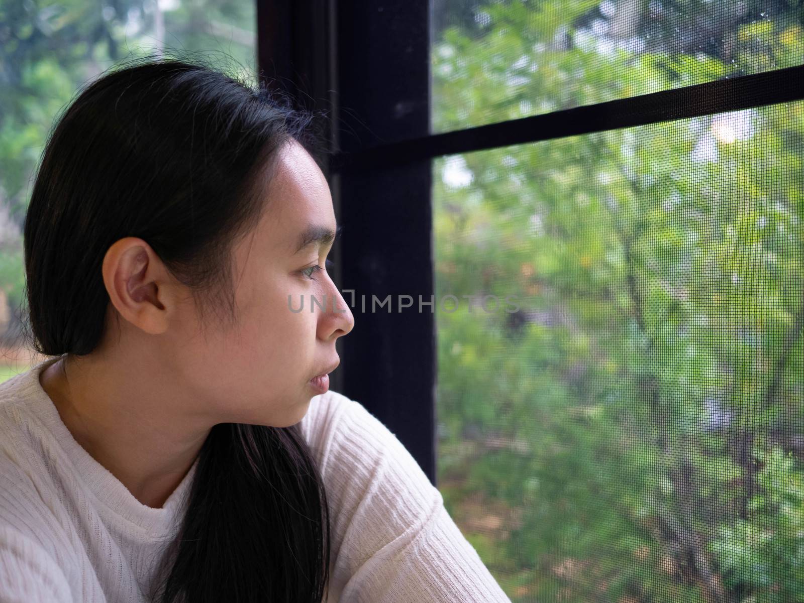 Portrait of Asian young sad woman looking through a window in a rainy day at home. by TEERASAK