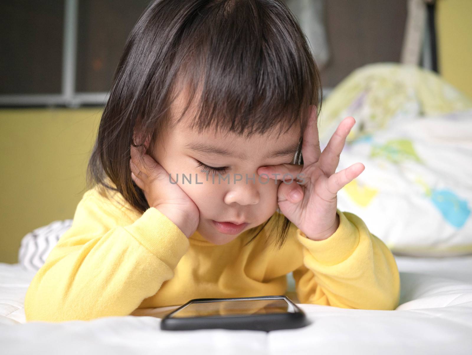 Asian little girl rubbing her eyes feel painful after watching smartphone on bed. Kid addicted to mobile phones.