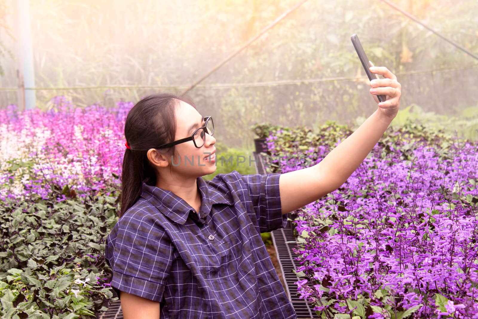Happy young woman making selfie by mobile phone with flowers plants in greenhouse.