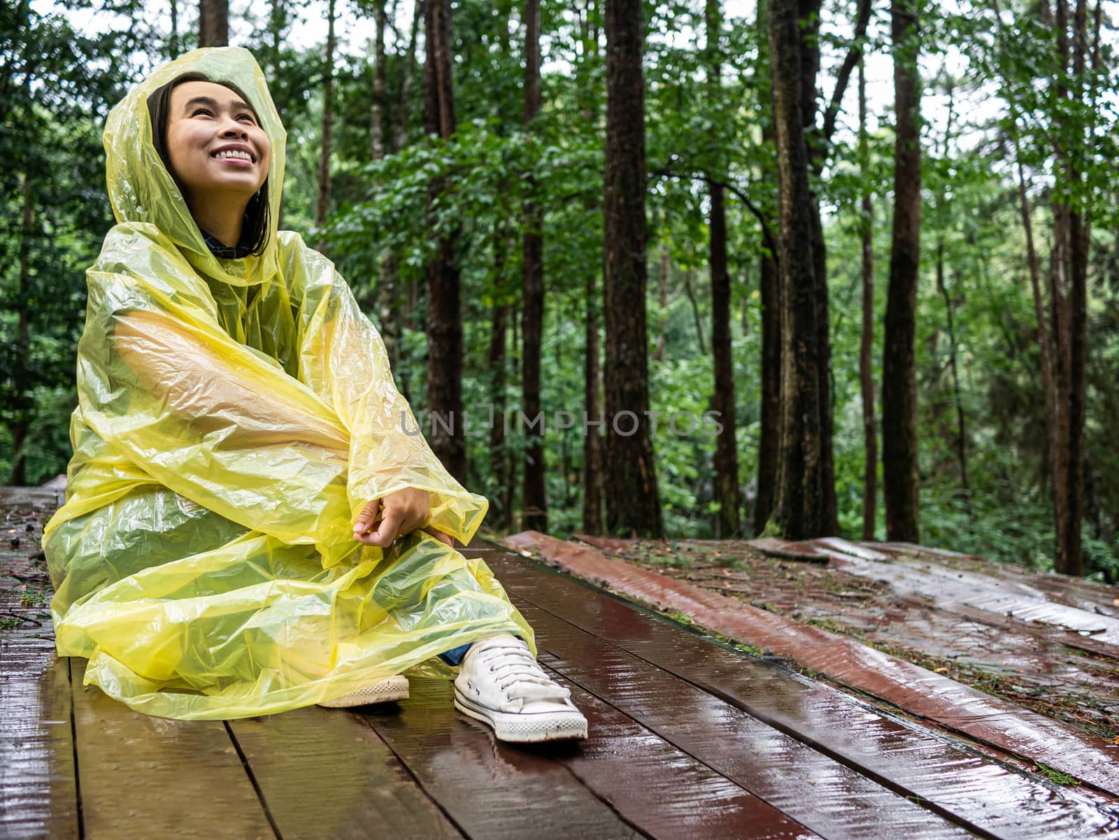 Happy Asian woman wearing yellow raincoat sit on wooden floor under rain. by TEERASAK