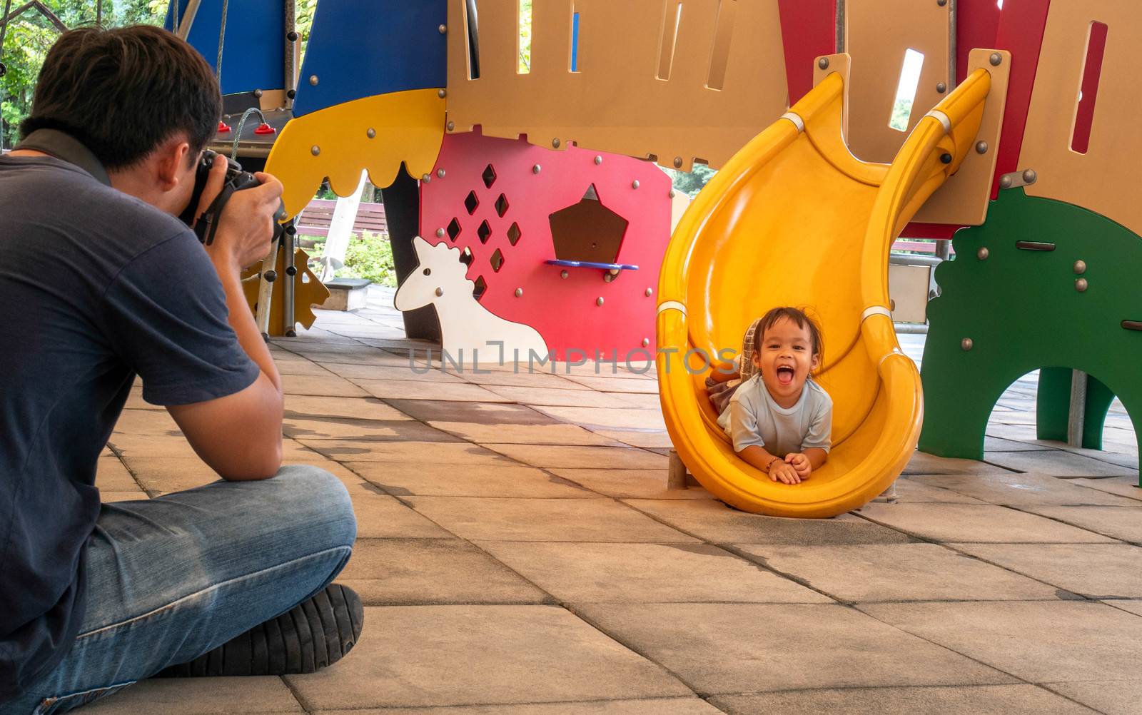 Father take a photo his daughter while having fun on slide at playground. by TEERASAK