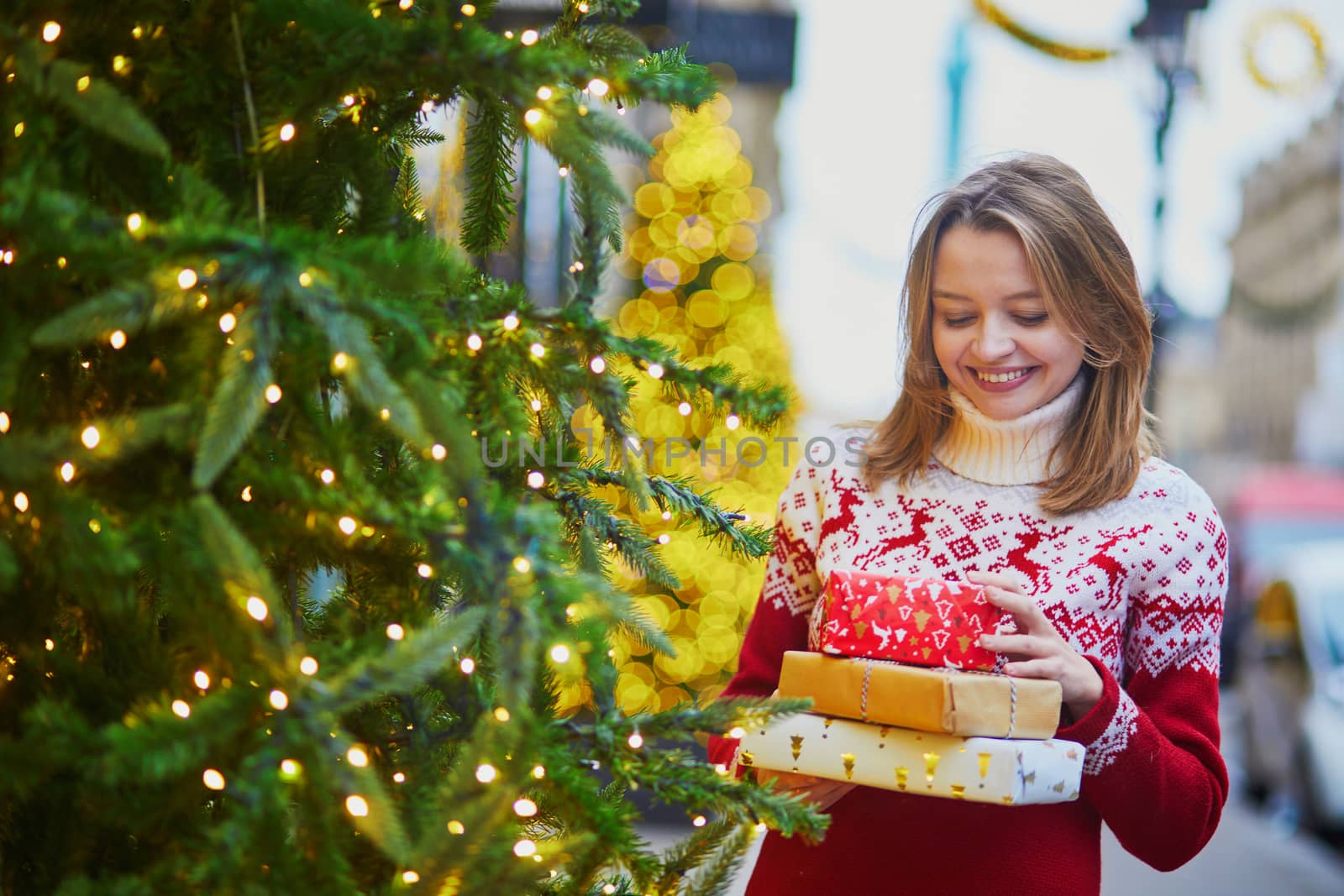 Happy young girl in warm red knitted sweater with pile of holiday gifts on a street of Paris decorated for Christmas