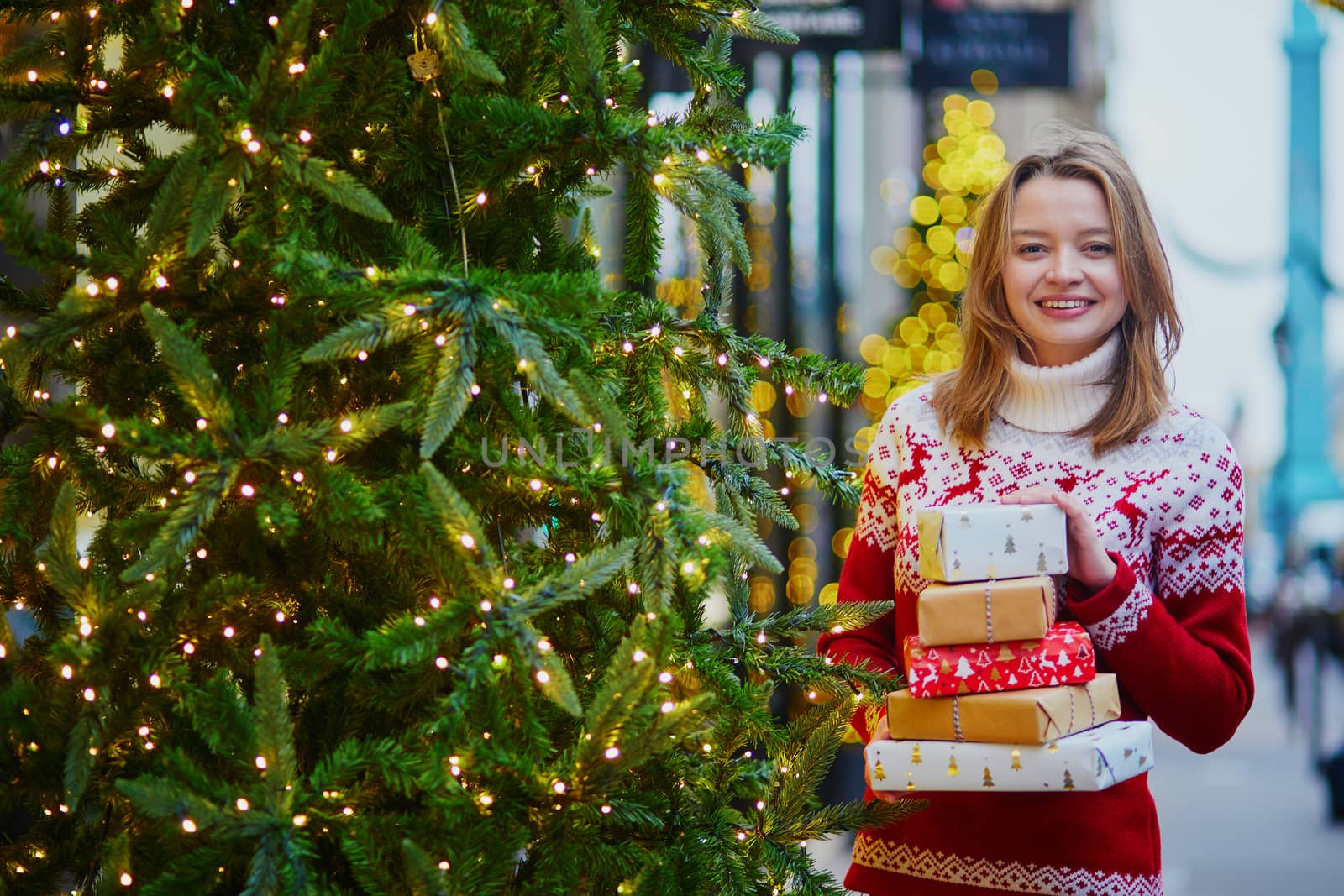 Happy young girl in warm red knitted sweater with pile of holiday gifts on a street of Paris decorated for Christmas