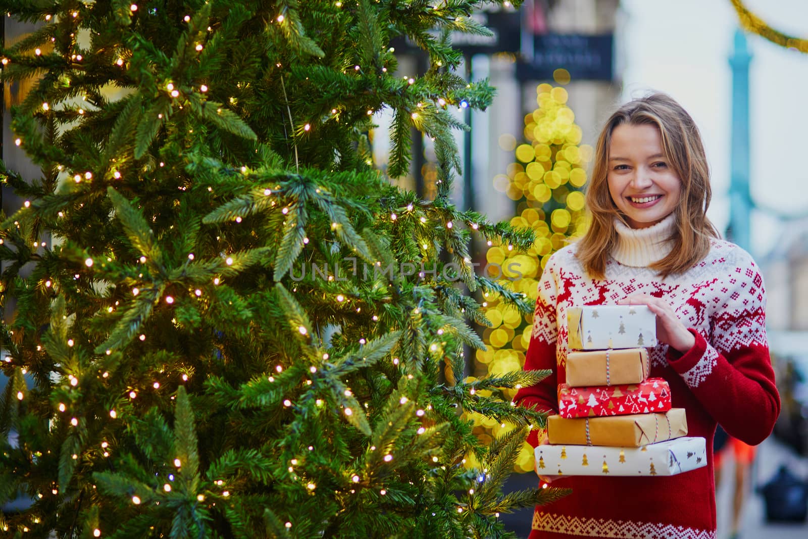 Happy young girl in holiday sweater with pile of holiday gifts on a street of Paris decorated for Christmas