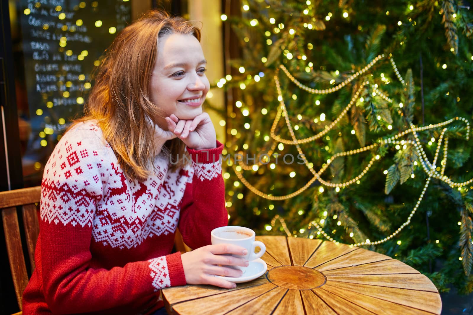 Cheerful young girl in holiday sweater drinking coffee or hot chocolate in cafe decorated for Christmas