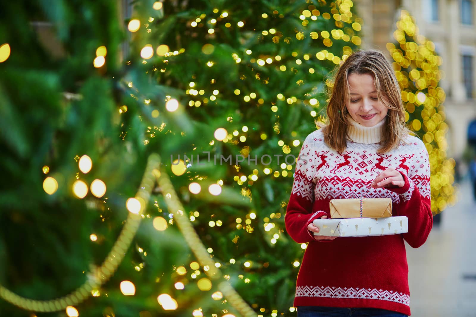 Happy young girl in holiday sweater with pile of holiday gifts on a street of Paris decorated for Christmas