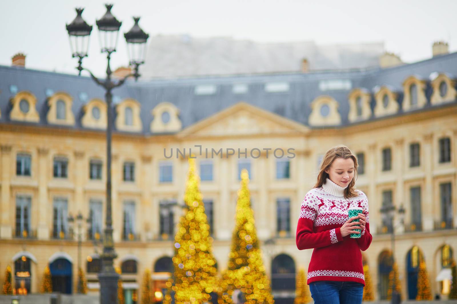 Happy young girl in holiday sweater walking with hot drink to go on a street of Paris decorated for Christmas