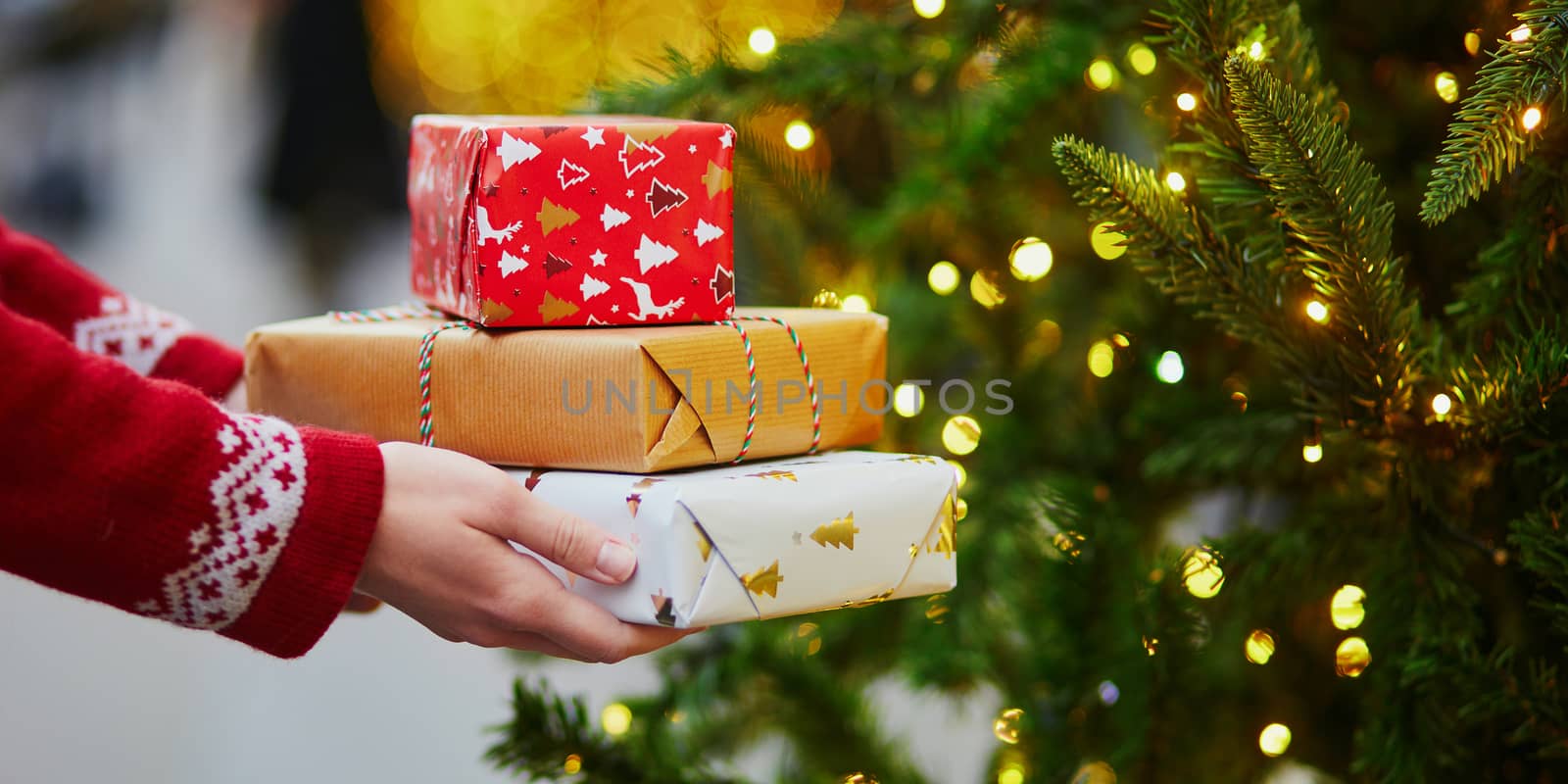 Woman hands holding pile of Christmas presents near New year tree decorated with lights and beads