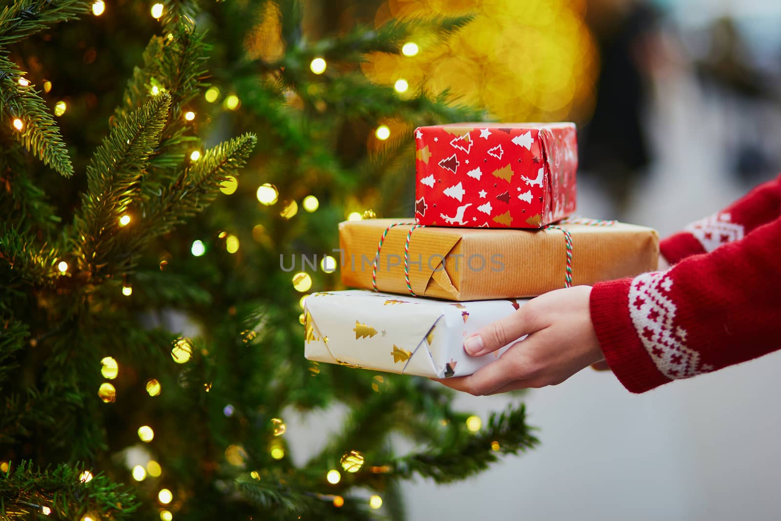 Woman hands holding pile of Christmas presents near New year tree decorated with lights and beads