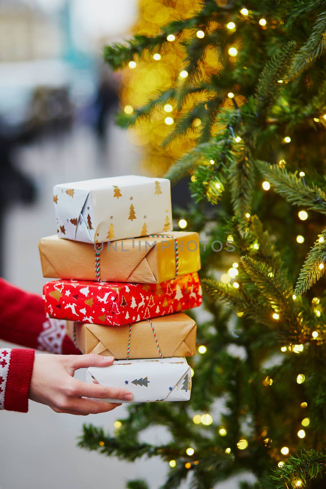 Woman hands holding pile of Christmas presents near New year tree decorated with lights and beads