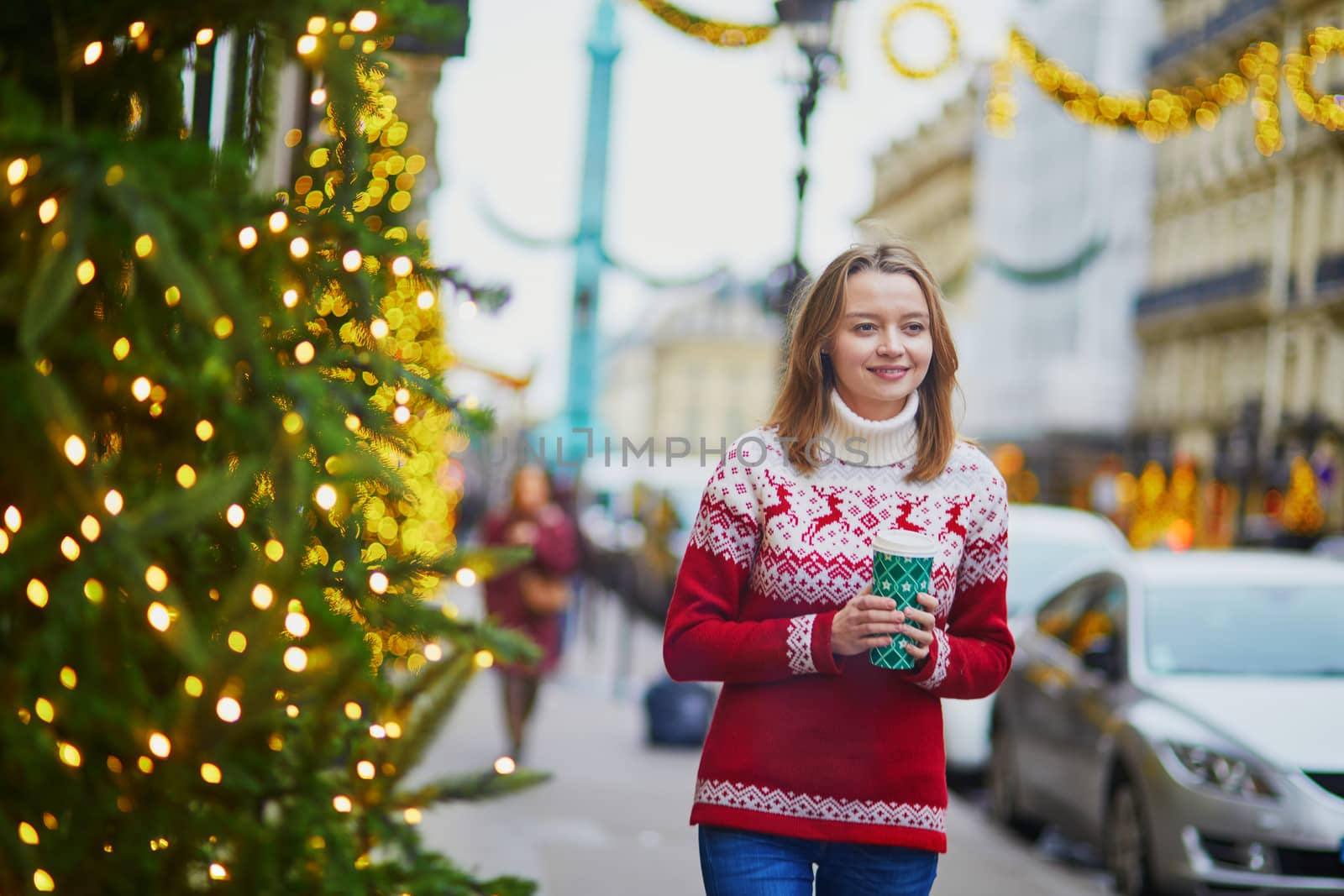 Happy young girl in holiday sweater walking with hot drink to go on a street of Paris decorated for Christmas