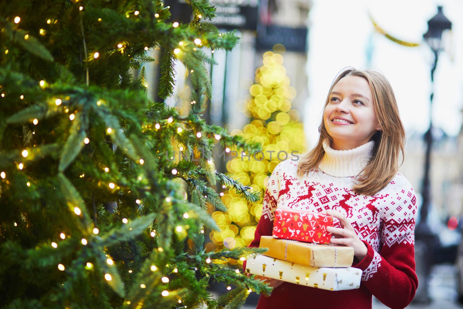 Happy young girl in warm red knitted sweater with pile of holiday gifts on a street of Paris decorated for Christmas