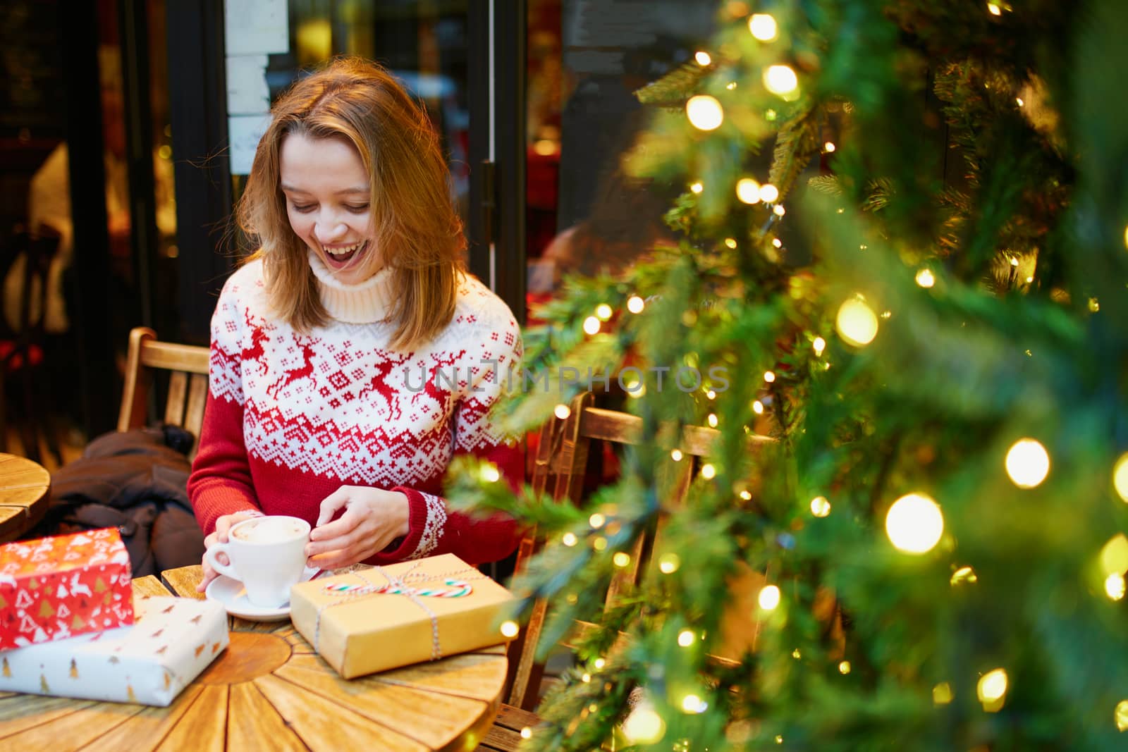 Cheerful young girl in warm red knitted holiday sweater drinking coffee or hot chocolate in cafe decorated for Christmas and unwrapping Christmas present