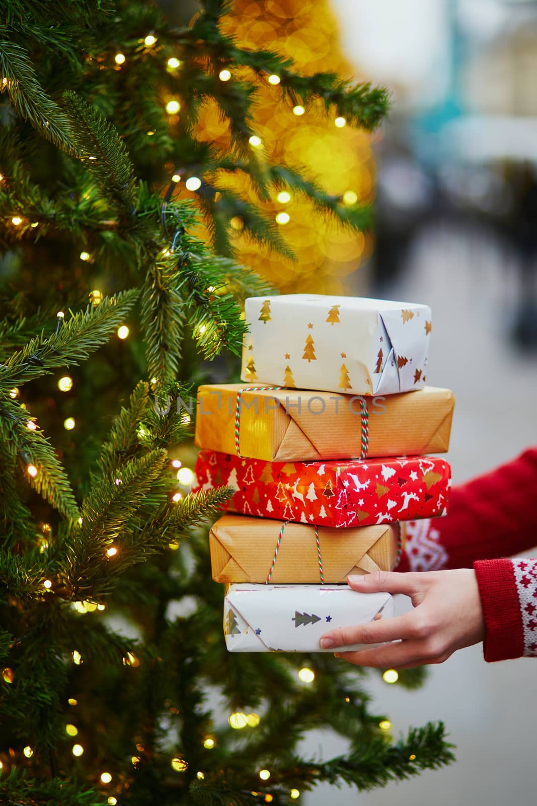 Woman hands holding pile of Christmas presents near New year tree decorated with lights and beads