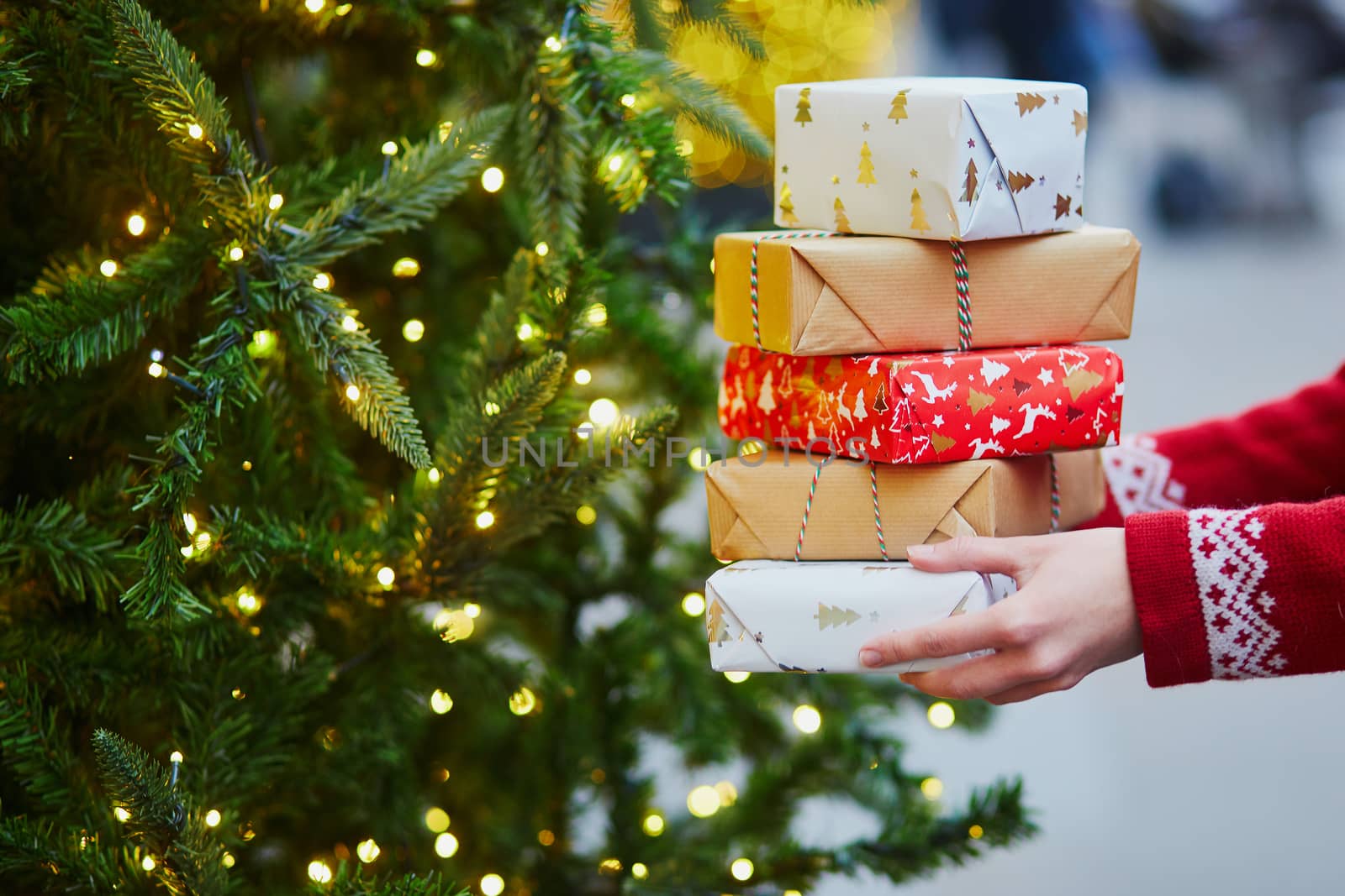 Woman hands holding pile of Christmas presents near New year tree decorated with lights and beads