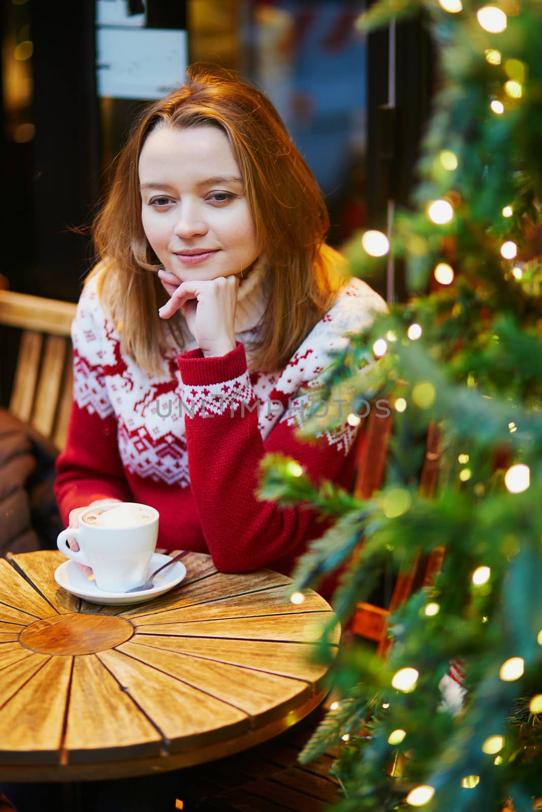 Cheerful young girl in warm red knitted holiday sweater drinking coffee or hot chocolate in cafe decorated for Christmas