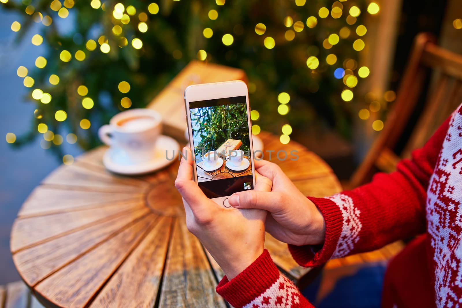 Woman taking photo of her coffee cup and Christmas present in cafe decorated for seasonal holidays