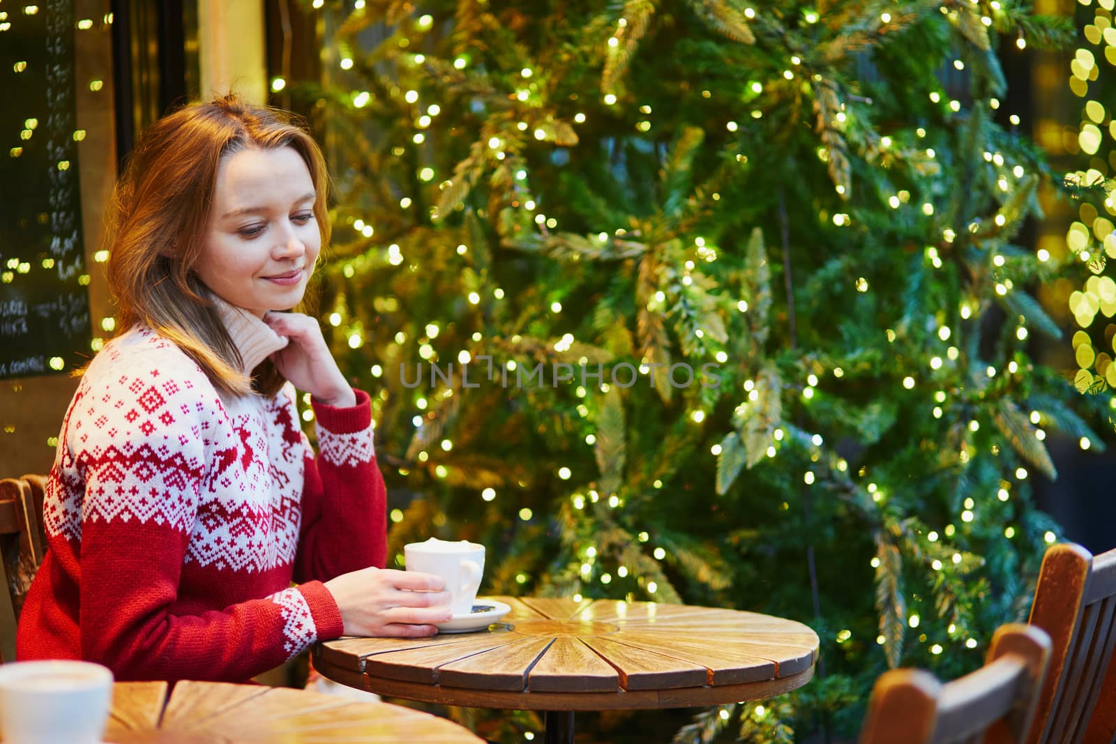 Cheerful young girl in holiday sweater drinking coffee or hot chocolate in cafe decorated for Christmas