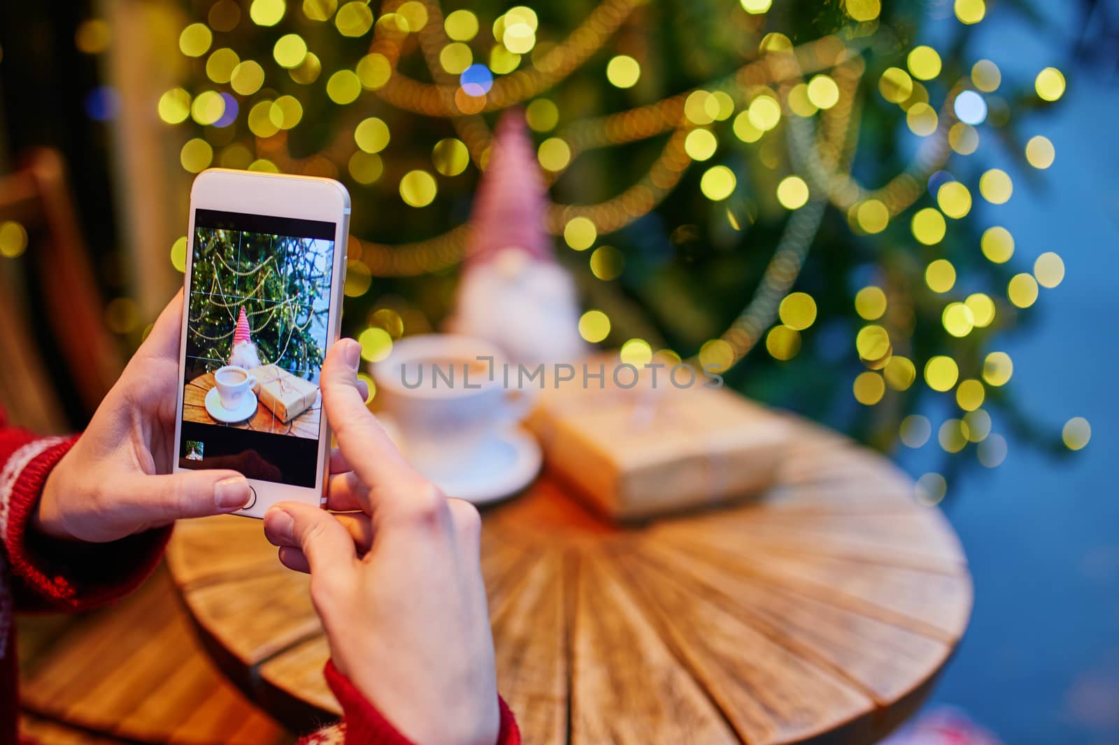 Woman taking photo of her coffee cup and Christmas present in cafe decorated for seasonal holidays