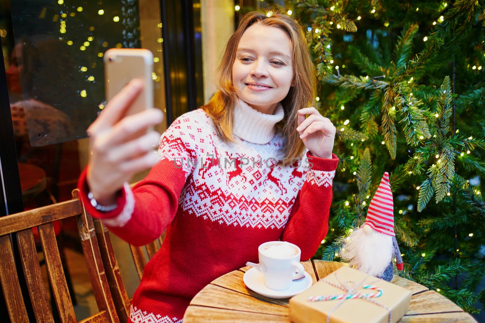 Cheerful young girl in warm red knitted holiday sweater in cafe decorated for Christmas, making selfie with her coffee cup and Christmas present