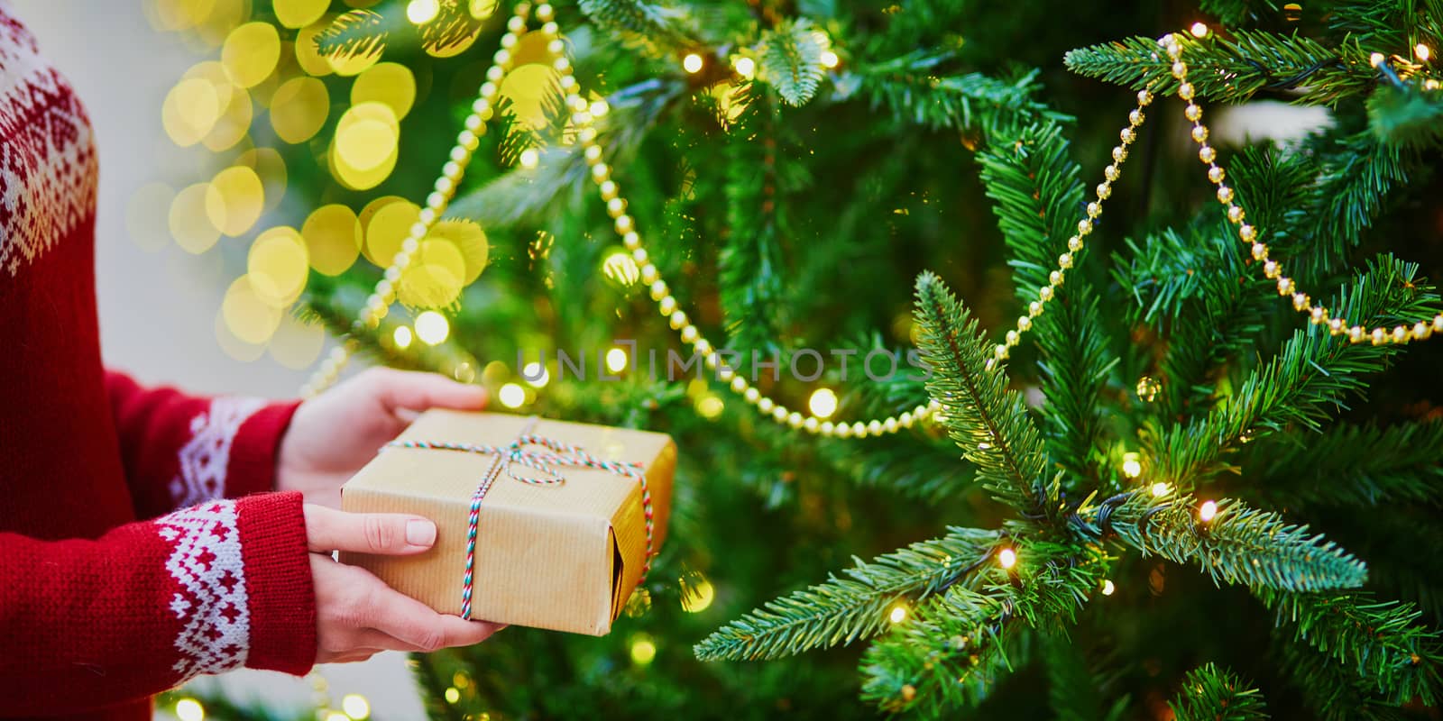 Woman hands holding Christmas present near New year tree decorated with lights and beads