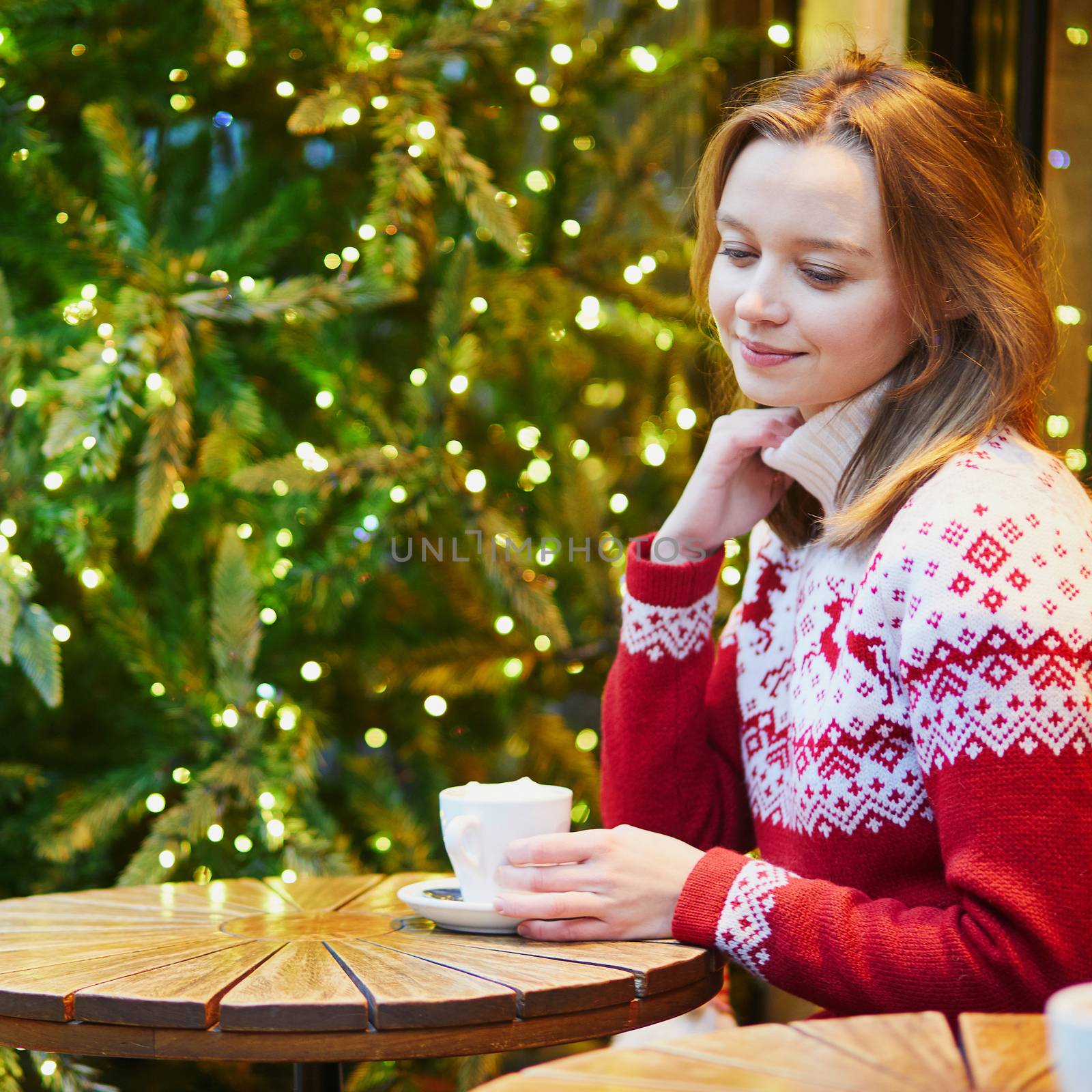 Cheerful young girl in warm red knitted holiday sweater drinking coffee or hot chocolate in cafe decorated for Christmas