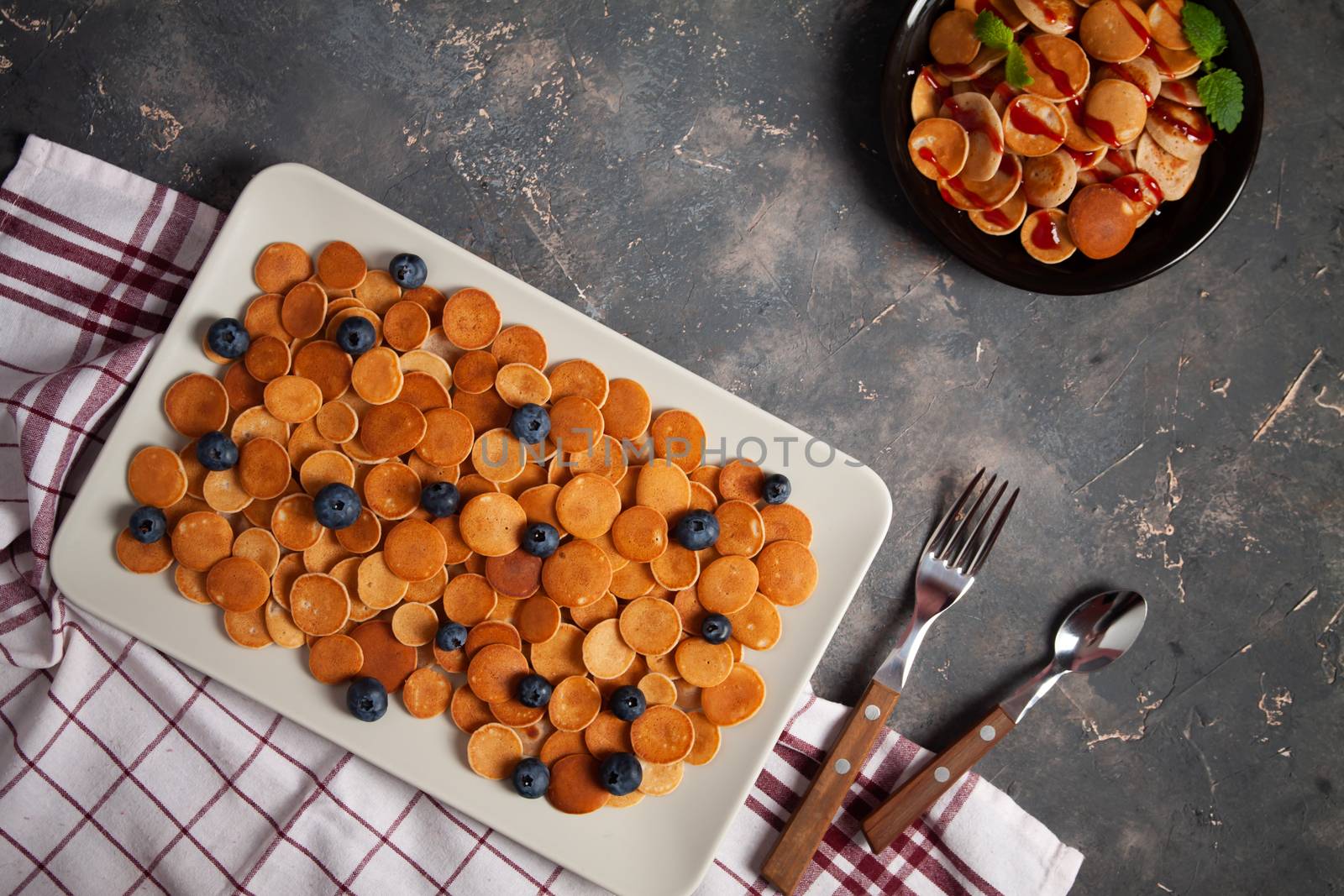 Trendy homemade breakfast, pancake cereal, mini pancakes on plates on dark concrete background with spoon and blueberry, peppermint with textile