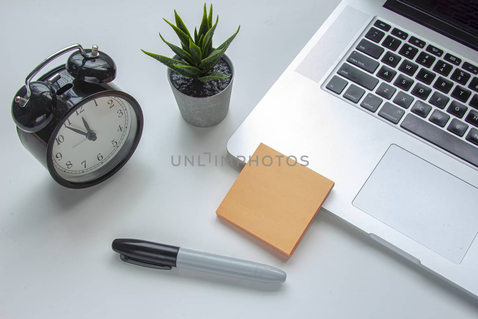 A note an a marker next to a laptop keyboard with a clock and a plant on a white table