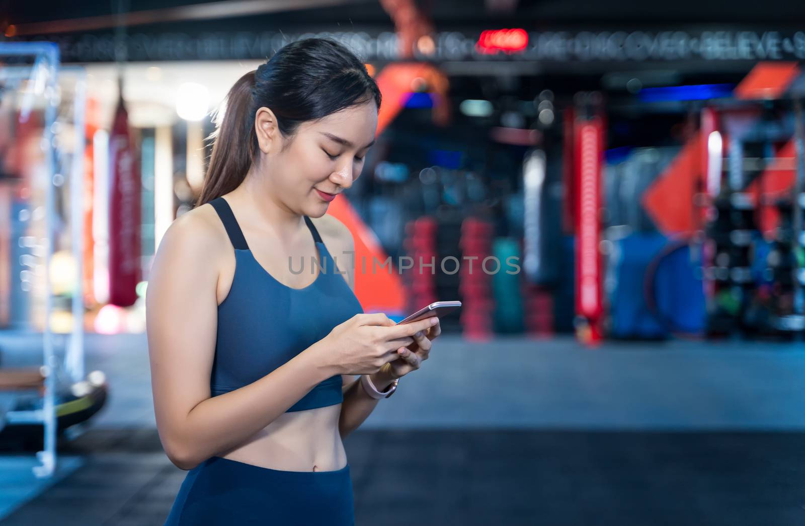 Asian Women are using mobile phones during exercise. She's standing, typing messages or chatting and smiling happily while staying during workouts with blur gym background and copy space