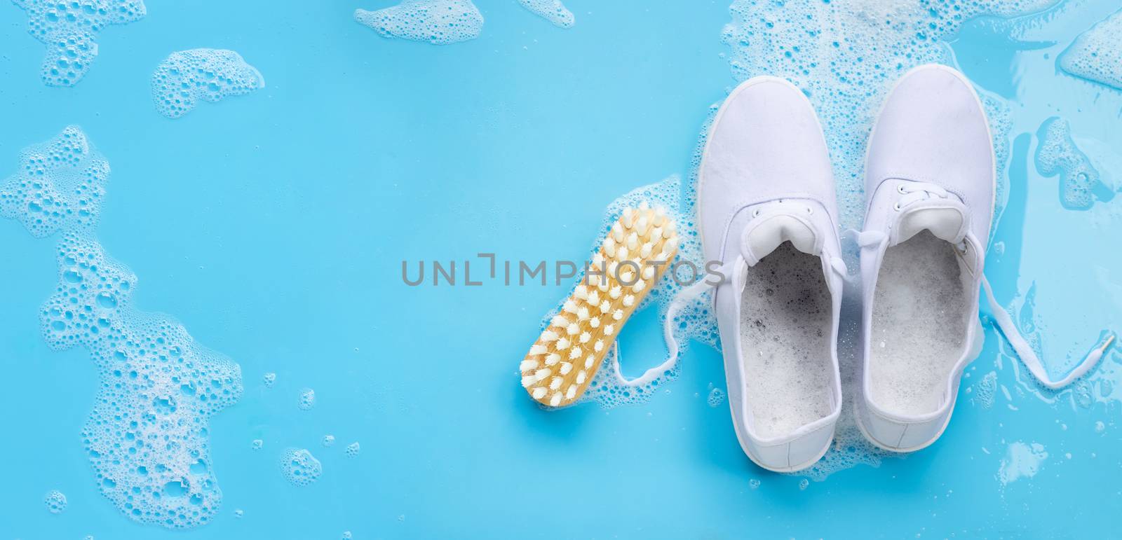 Sneakers with foam of powder detergent water dissolution and wooden brush on blue background. Washing dirty shoes. Top view