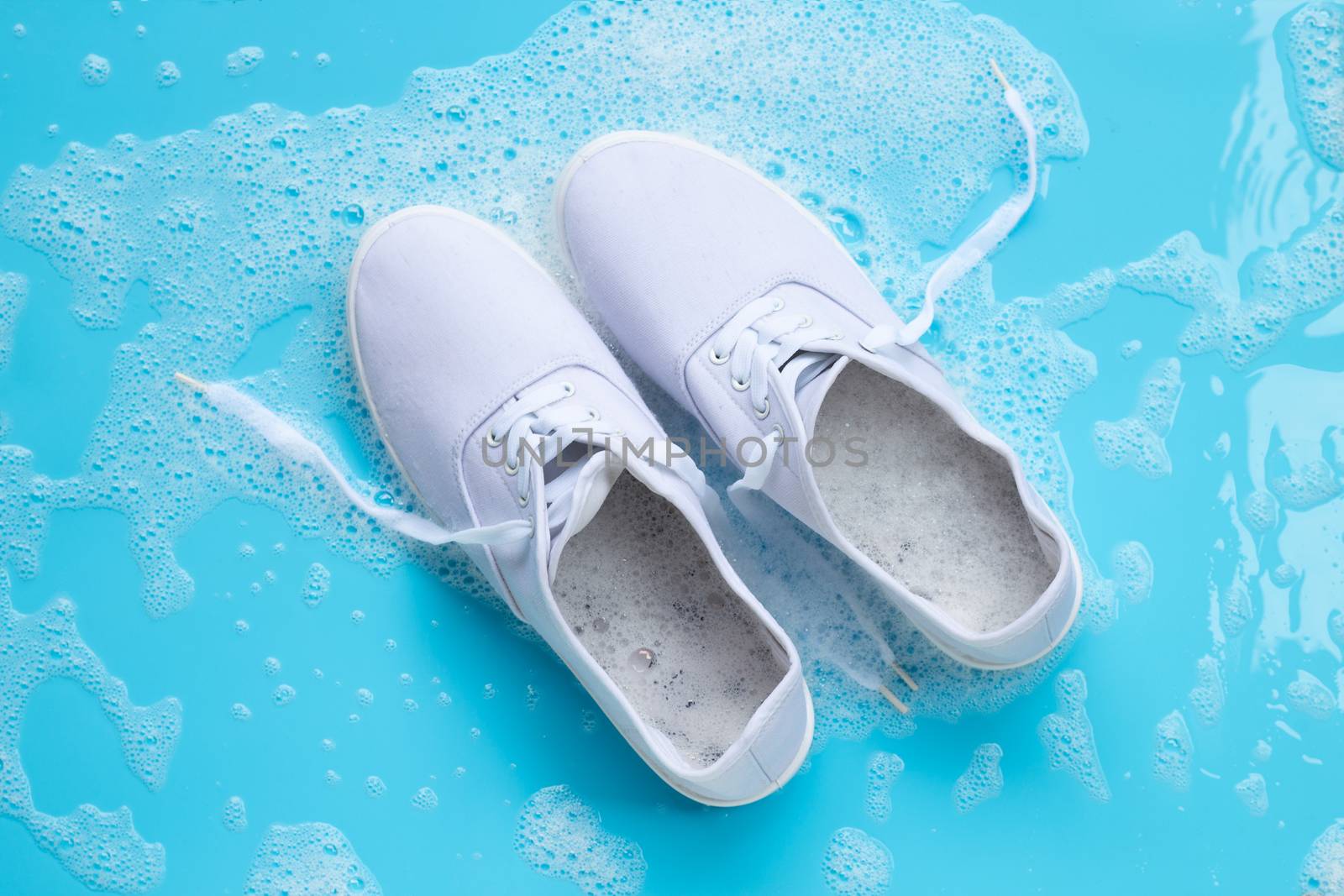 Sneakers with foam of powder detergent water dissolution on blue background. Washing dirty shoes. Top view