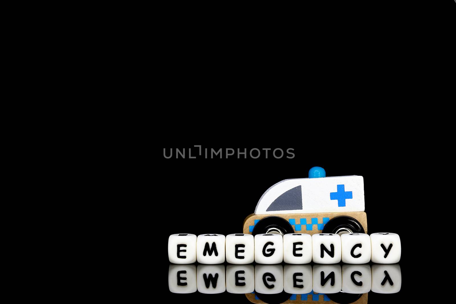 A wooden toy ambulance and white alphabet letters spelling the word emergency, rapid and prompt medical attention concept.
