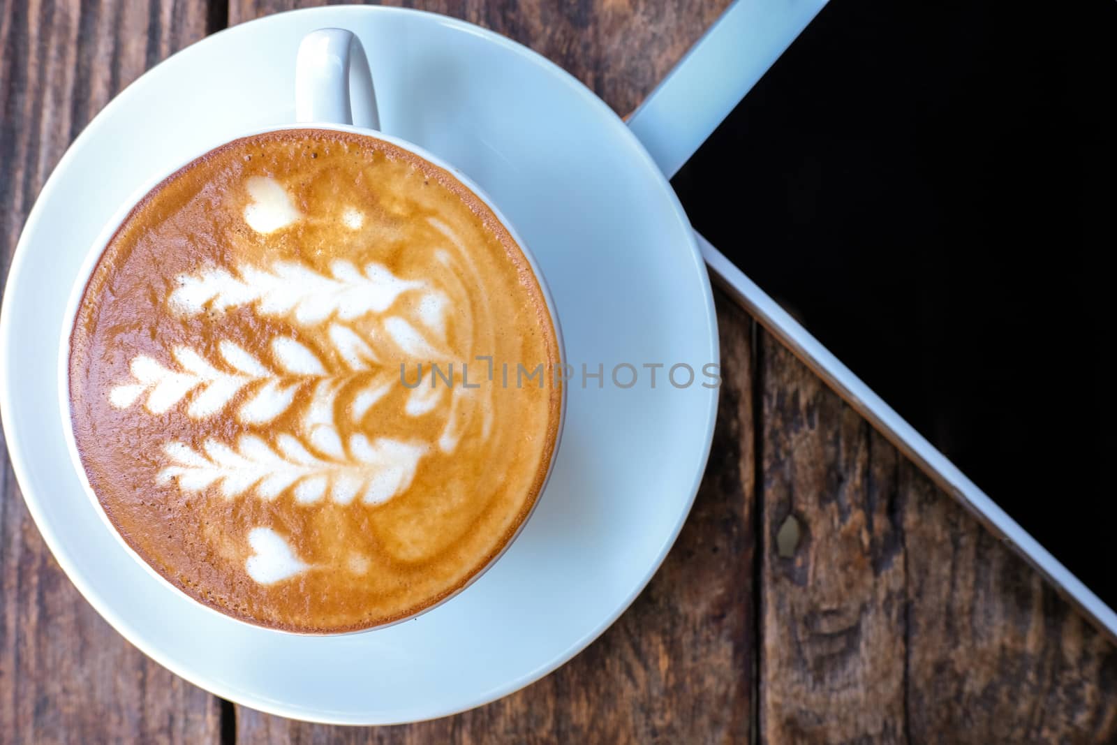 a cup of hot cappuccino coffee with latte art pattern and a tablet on a brown wooden table