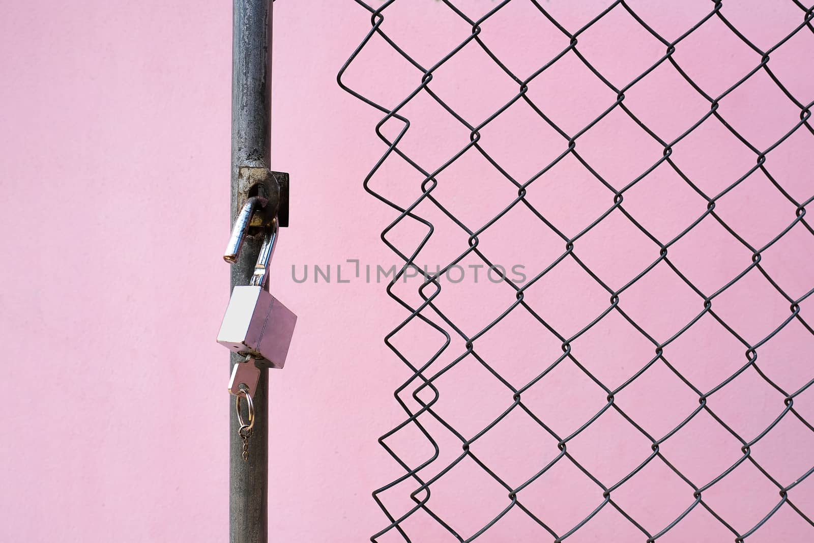 an old rusty metal padlock and a key at an opened wired gate in front of a pink colored concrete wall, security concepts