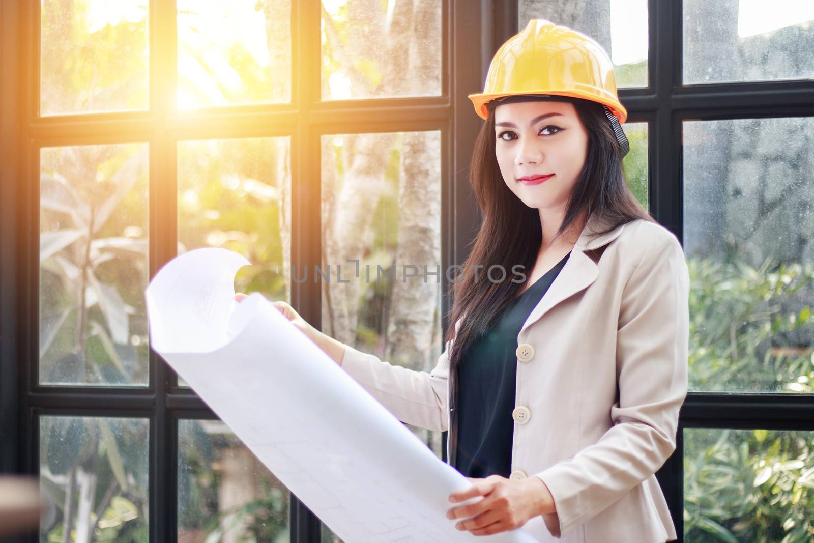 portrait of beautiful Asian woman architect builder with yellow helmet hard hat studying blueprint plan of the rooms, serious civil engineer working with documents on construction site
