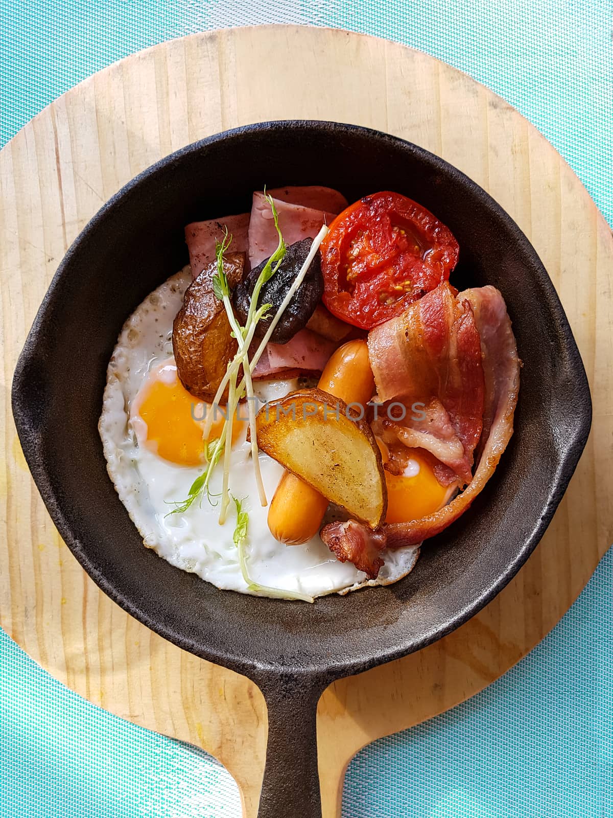 Breakfast in cooking pan with fried eggs, sausages, bacon , tomato , potato and vegetable on wood block background on the table in the kitchen. morning light effect by asiandelight